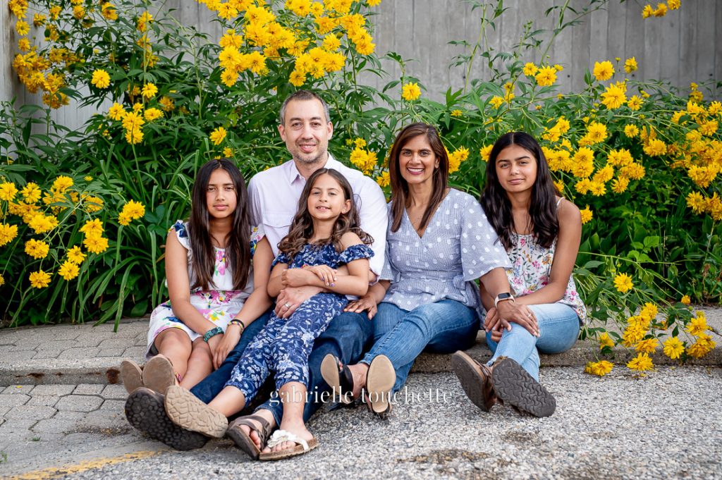 A family sitting on the ground in front of some bright yellow flowers and a concrete wall in Winnipeg