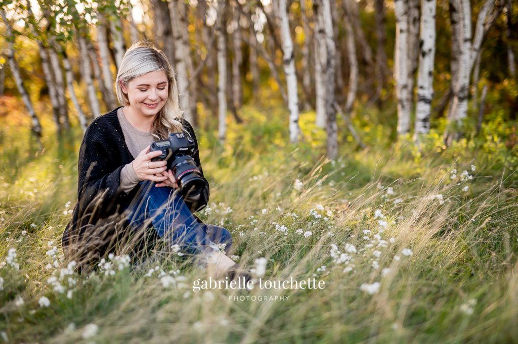 A photographer sitting in tall grass with wildflowers and birch trees in the background for a portrait