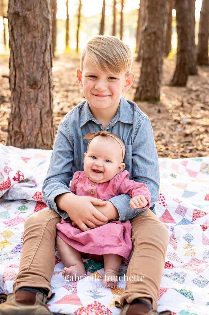 A big brother holding his baby sister in his arms on a homemade quilted blanket on the forest floor