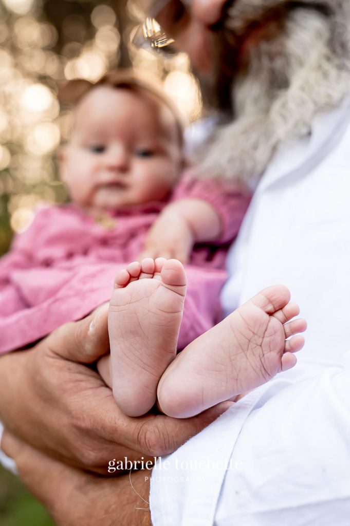 A macro photo of a baby's foot and toes being cradled in her fathers arms wearing a pink dress with bokkeh light in the background