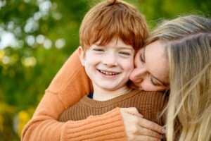 Candid family photo of a mother hugging and kissing her smiling son during a relaxed outdoor session in Winnipeg, capturing a natural moment of connection.