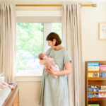 at home newborn photo session with mom holding baby girl in front of the window in the nursery