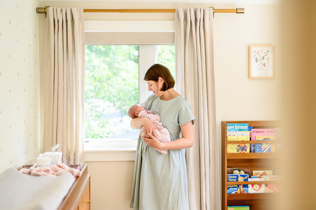 at home newborn photo session with mom holding baby girl in front of the window in the nursery